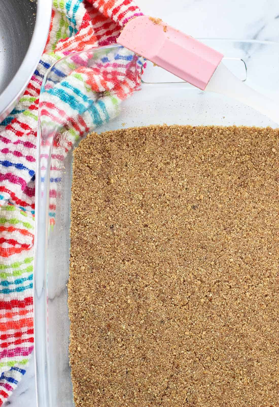 The crust mixture pressed into the bottom of a rectangular glass pan with a silicone spatula on top