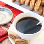 Two biscotti accompanying a cup of coffee next to the full tray of cookies.