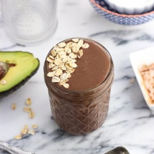 A smoothie in a glass jar topped with oats beside an avocado half and an empty measuring cup and bowls