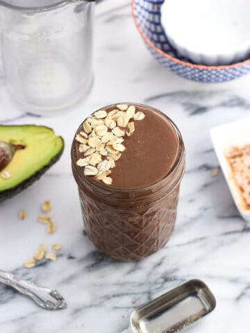 A smoothie in a glass jar topped with oats beside an avocado half and an empty measuring cup and bowls