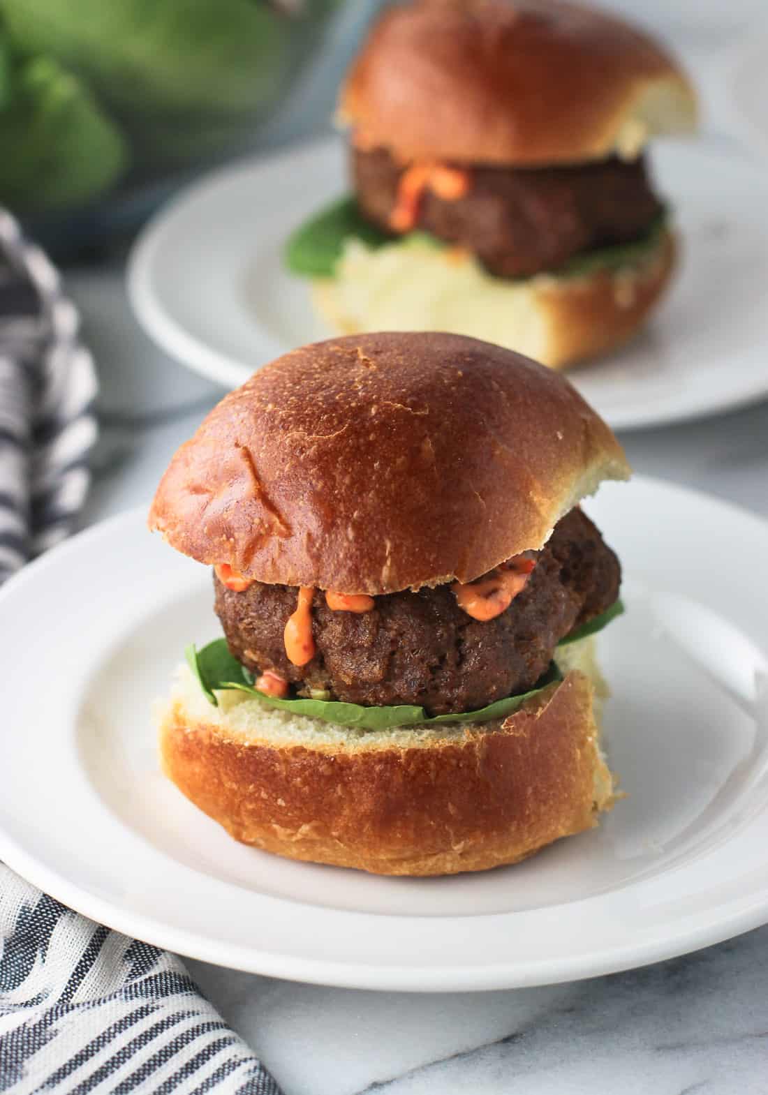 A turkey burger slider, with lettuce and chili garlic sauce, on a small slider bun on a plate with another in the background