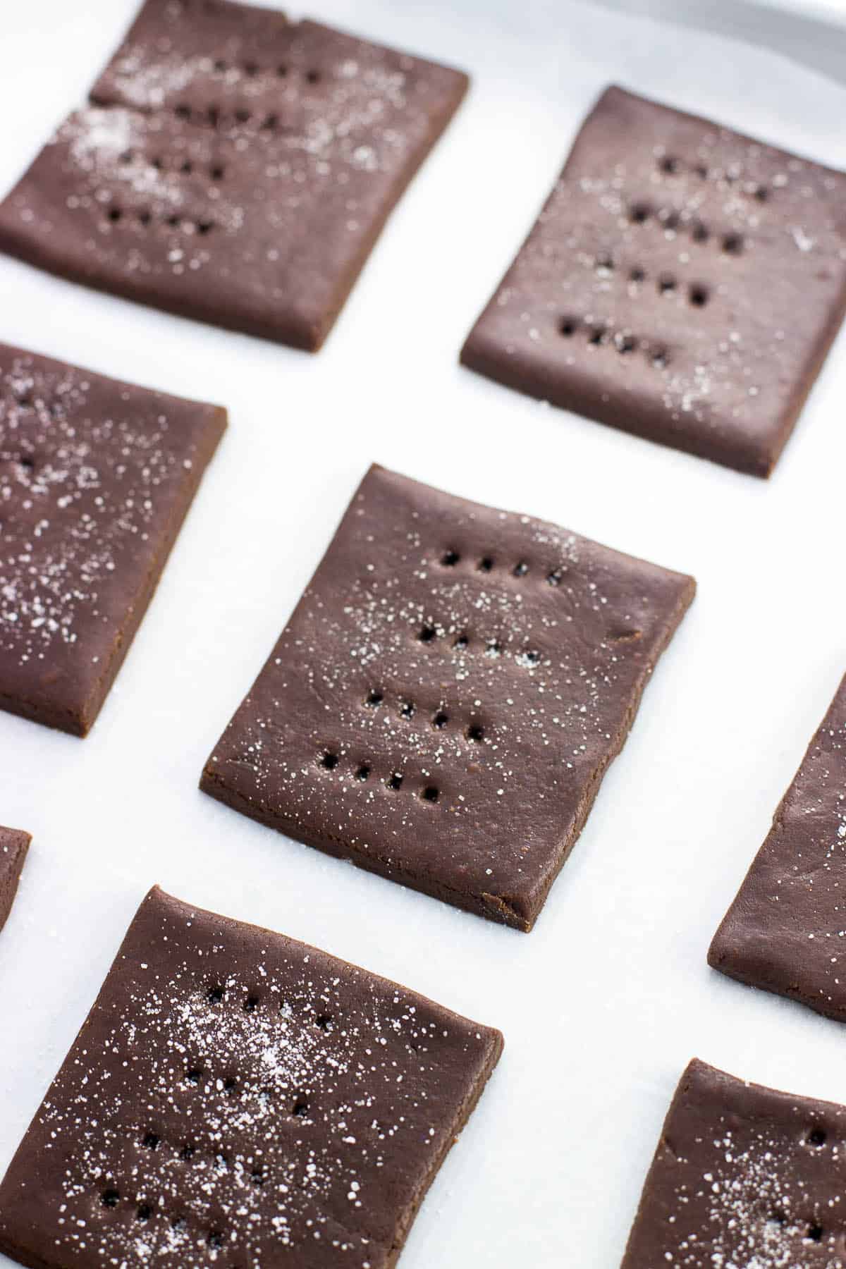 Squares of chocolate graham dough dusted with sugar on a parchment-lined baking sheet before going in the oven.