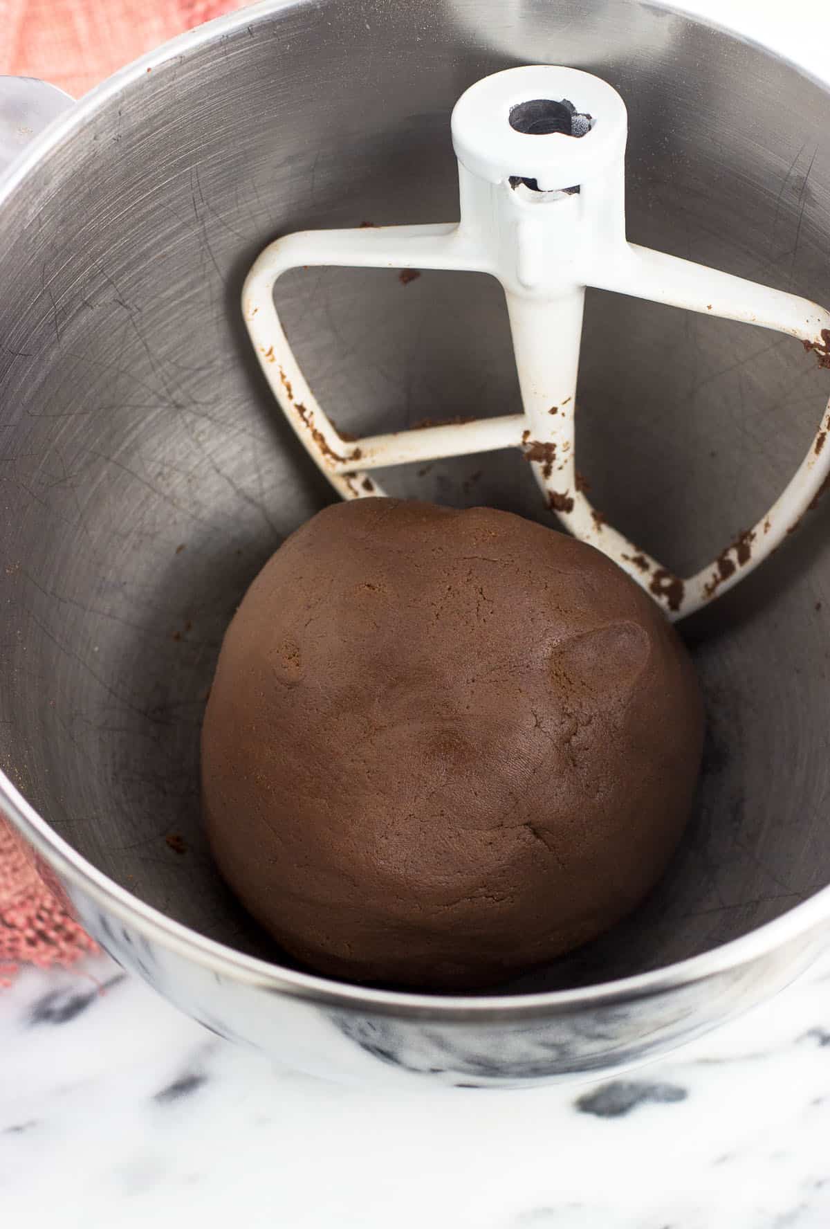 A ball of chocolate dough in the bowl of a stand mixer next to the paddle attachment