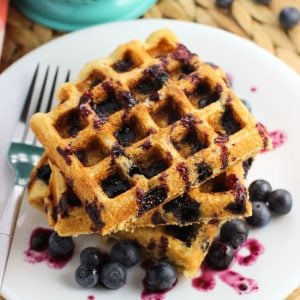 Three waffles on a plate with fresh blueberries and drizzled with blueberry maple syrup