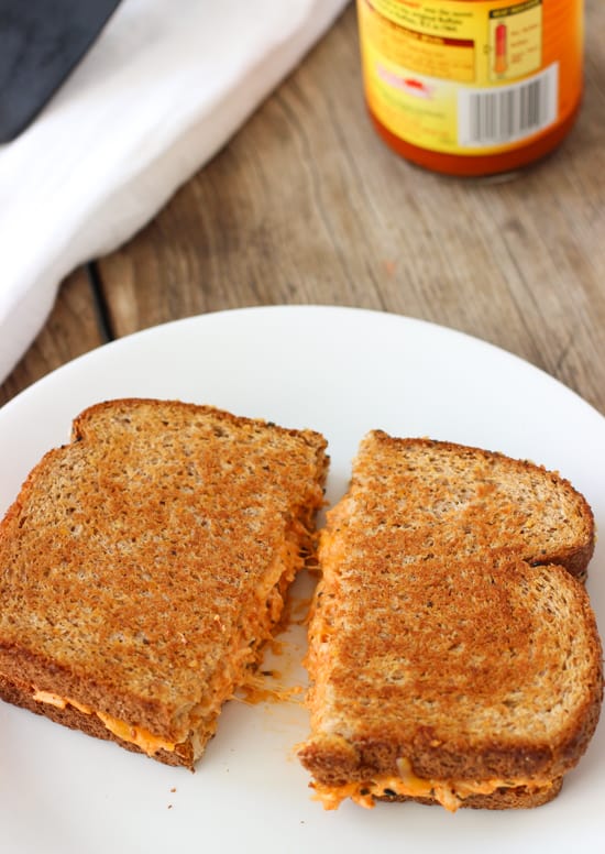 A grilled cheese sandwich on a plate with a bottle of buffalo sauce in the background