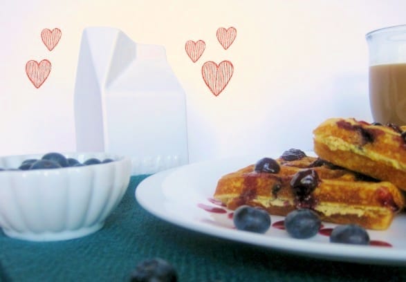Waffles and blueberries on a plate next to a ceramic carton of milk and blueberries.