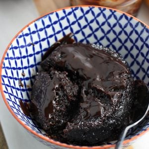 A mug cake turned out into a small bowl with a spoon and topped with hot fudge