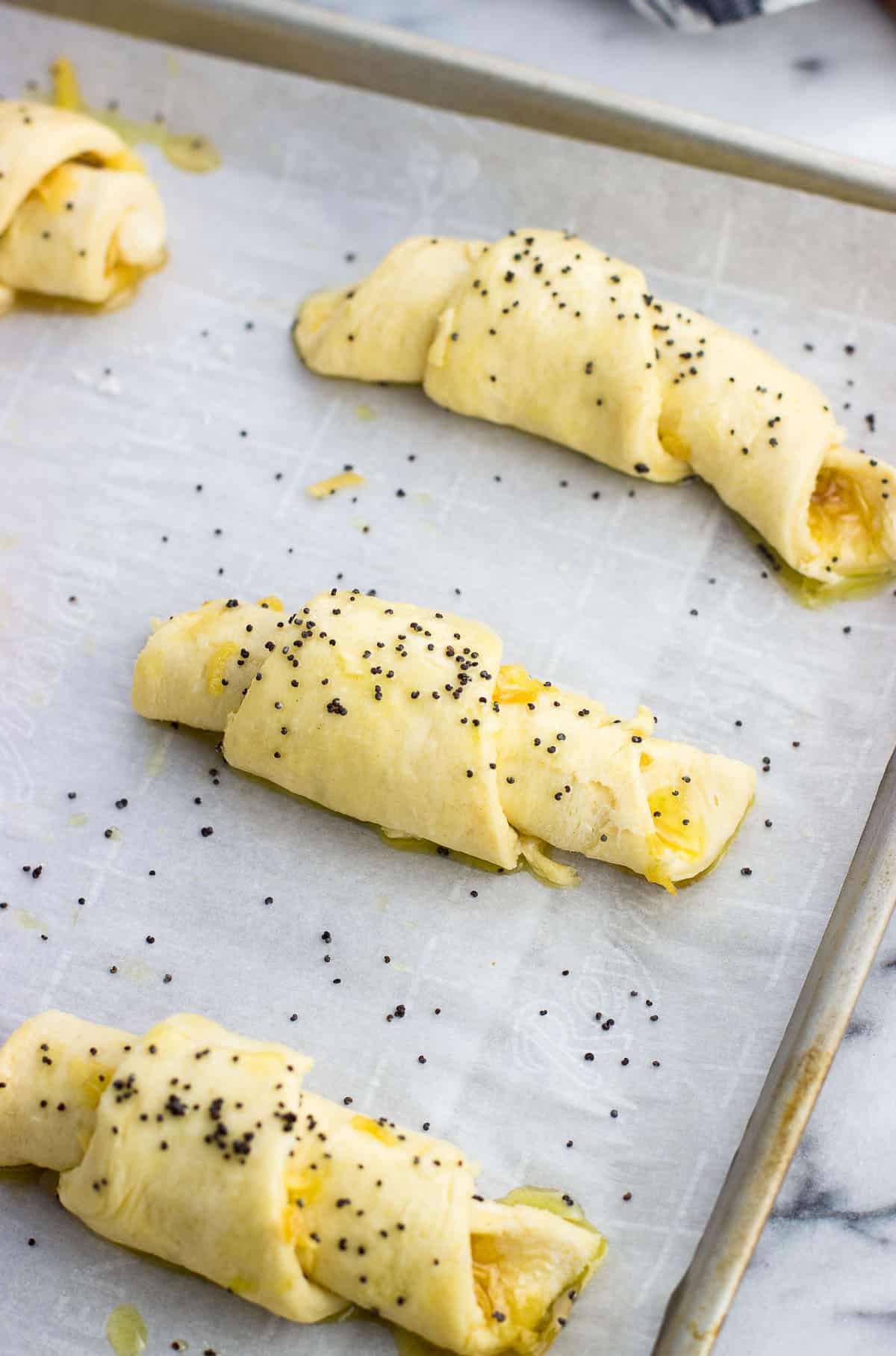 Rolled up crescent rolls topped with a brush of oil and poppy seeds on a baking sheet before being baked