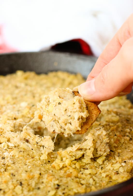 A hand lifting a dip-scooped cracker out of the pan of dip.