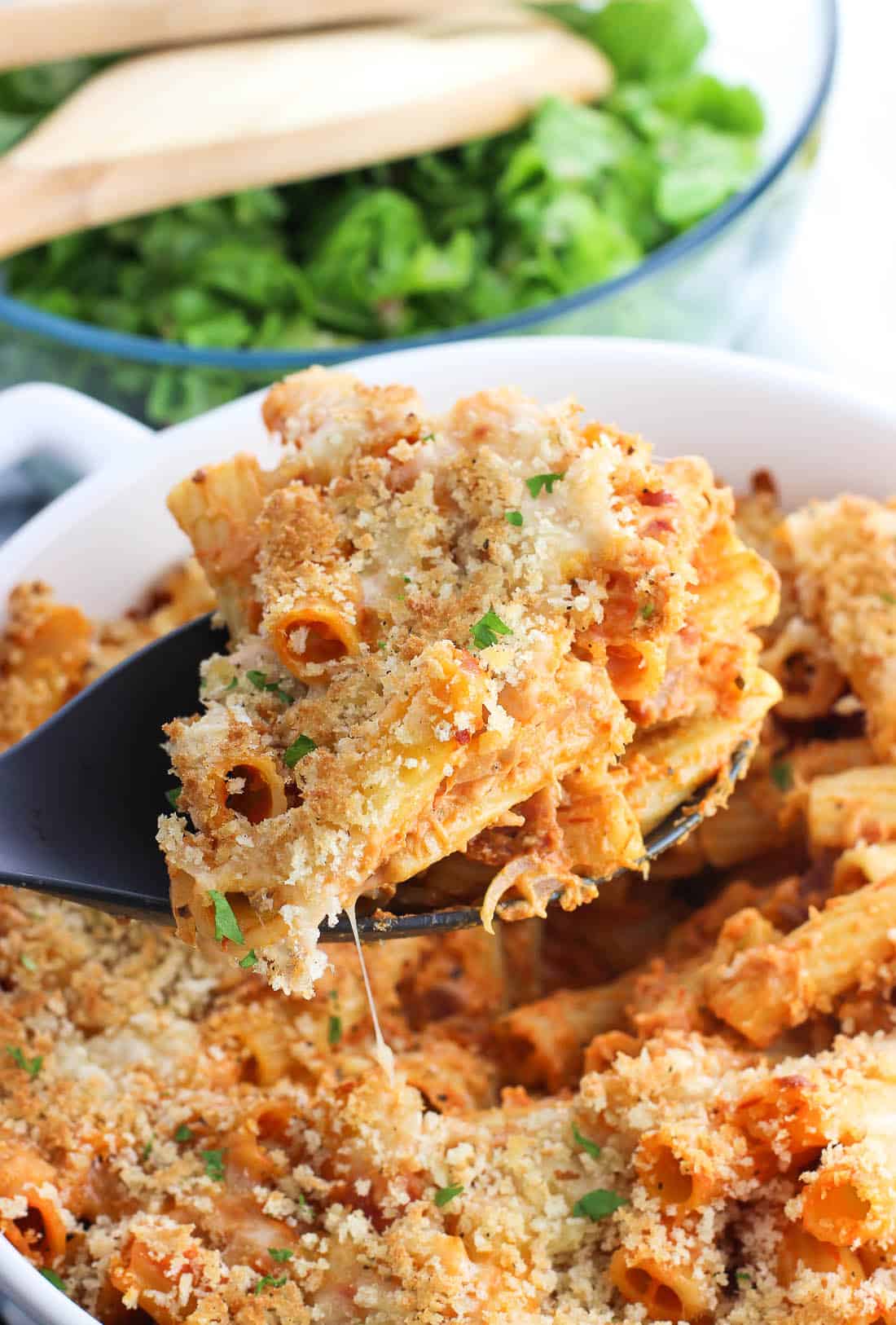 A serving spoon lifting out a scoop of baked pasta from the baking dish.