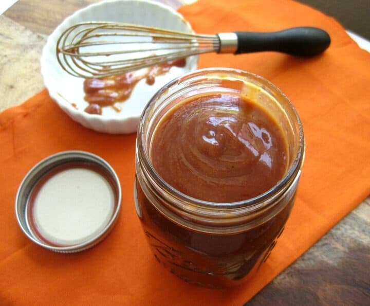 A glass jar of BBQ sauce next to a whisk.