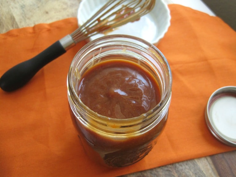 A glass jar of BBQ sauce next to a whisk.