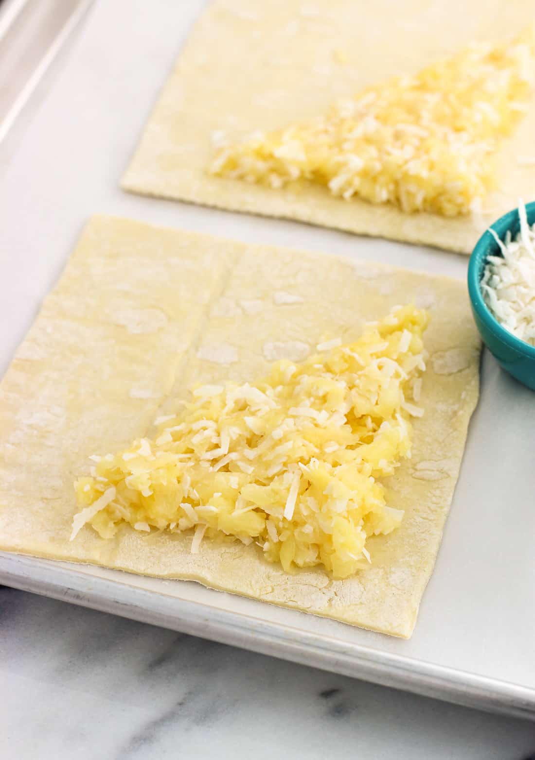 Puff pastry squares on a parchment-lined baking sheet, half filled (on the diagonal) with crushed pineapple and flaked coconut