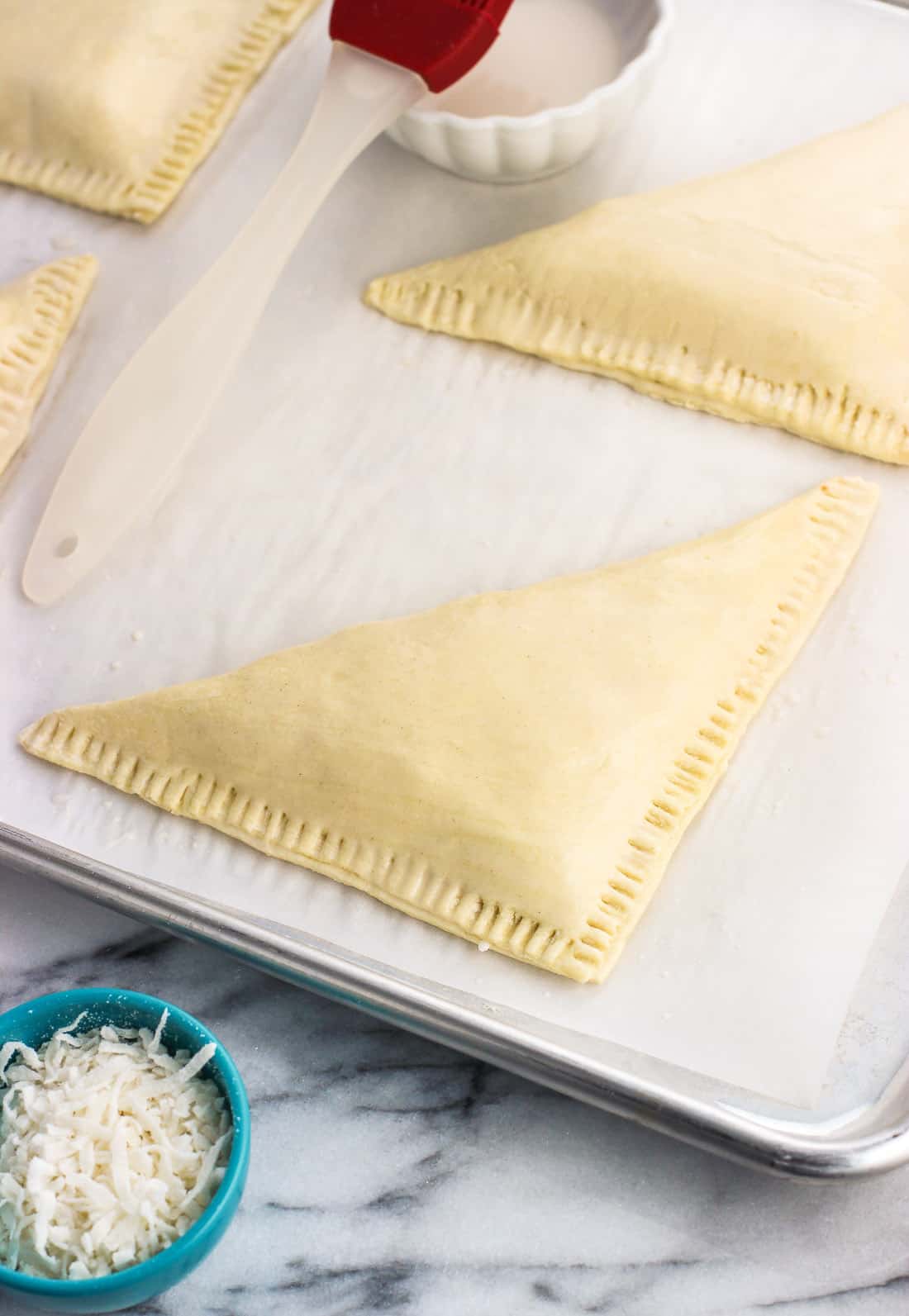 Triangular turnovers with two edges crimped on a parchment-lined baking sheet before going in the oven