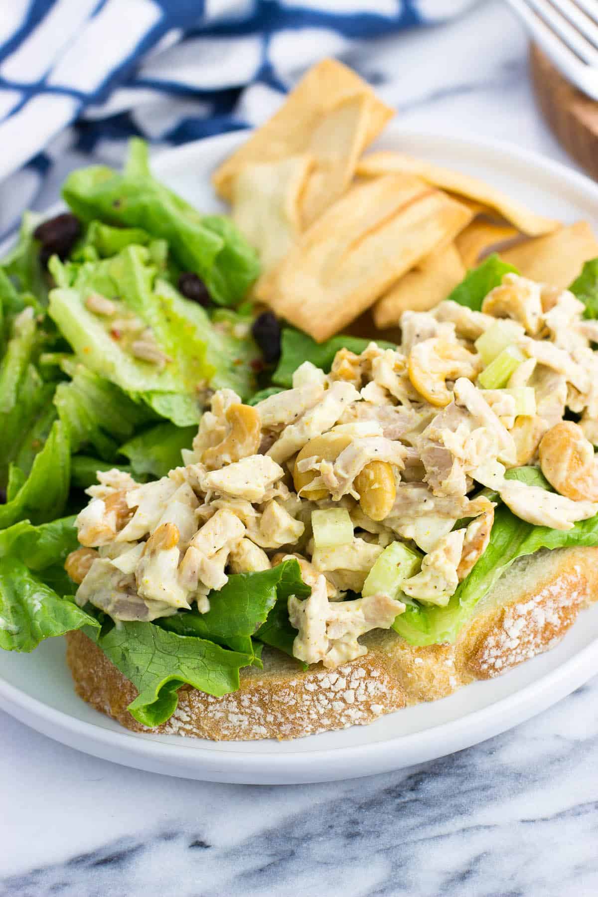 An open-face chicken salad sandwich on a plate with a side salad and pita chips