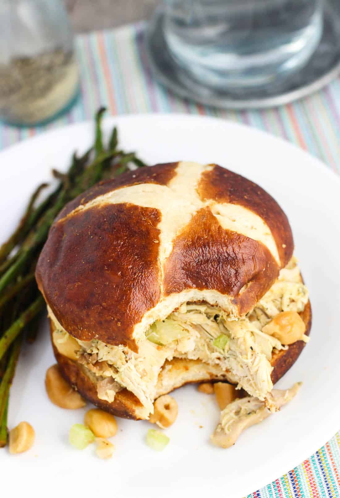 Chicken salad on a pretzel bun with a bite taken out of it. The sandwich is on a plate with sautéed asparagus.