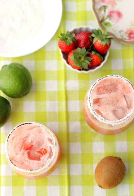 Two cocktails, fresh strawberries, limes, and a kiwi on a tablecloth.