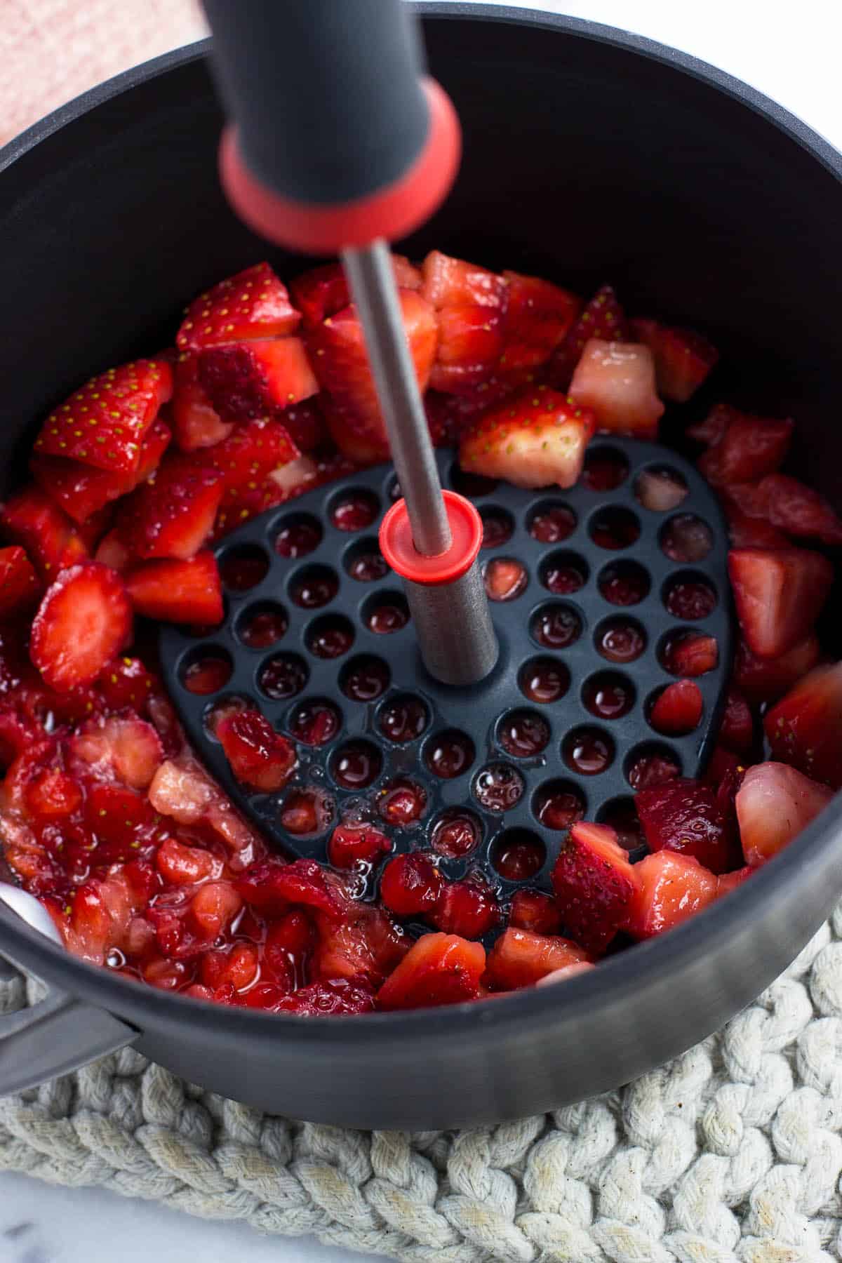 A potato masher being pressed into the chopped strawberries in a saucepan.