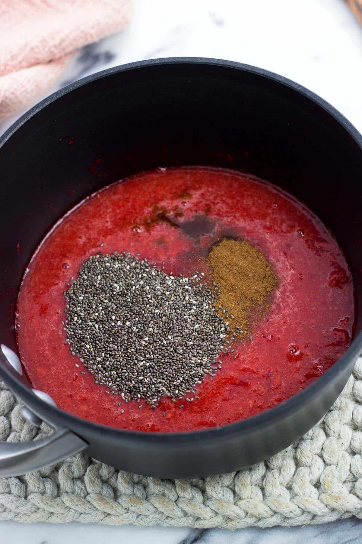 Mashed strawberry puree in a saucepan with chia seeds, cinnamon, and vanilla extract poured in.