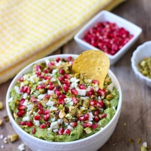 A tortilla chip in a bowl of guacamole next to little bowls of pomegranate arils and pistachios.