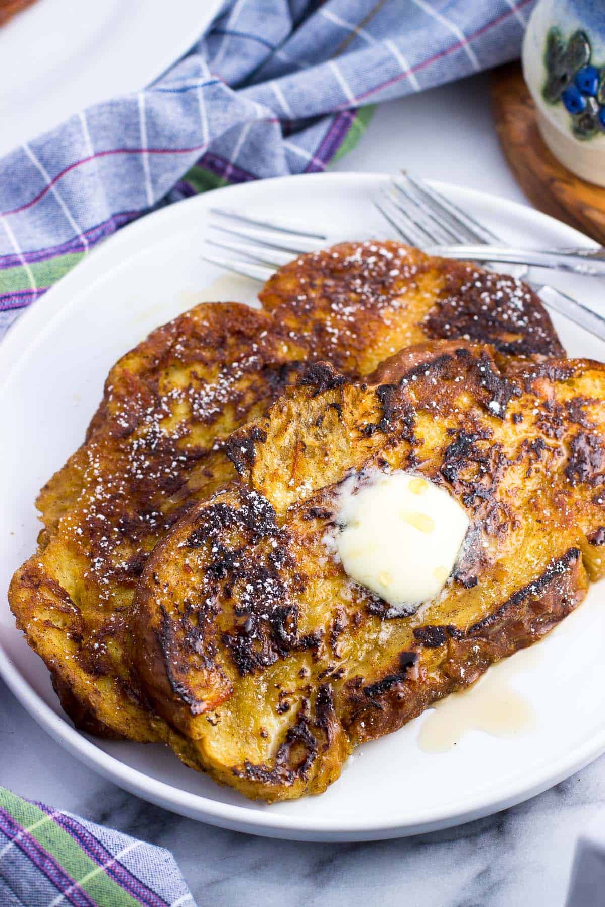 Two slices of challah french toast on a plate with forks, maple syrup, and butter.