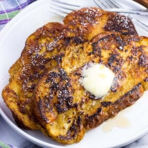 Two slices of challah french toast on a plate with maple syrup, a pat of butter, and forks.