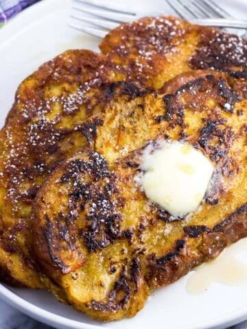Two slices of challah french toast on a plate with maple syrup, a pat of butter, and forks.
