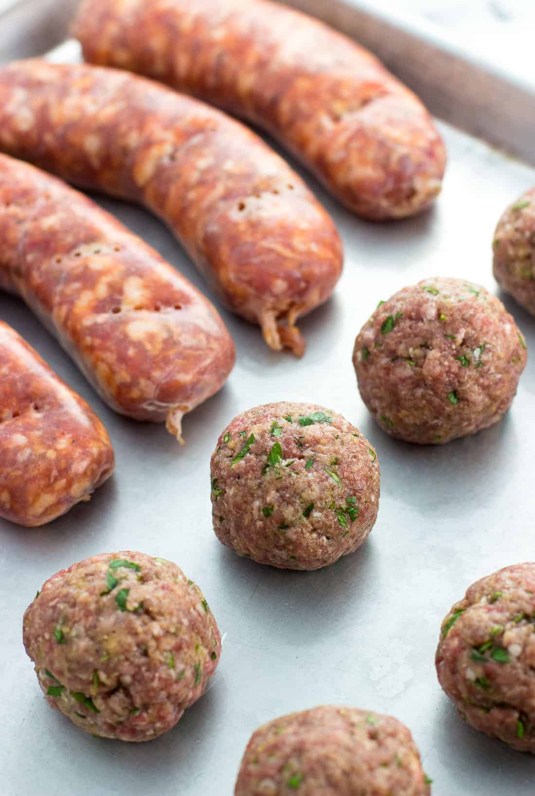 Meatballs and sausage links on a large metal baking sheet before baking in the oven