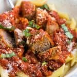A bowl of Sunday sauce with meatballs and sausage served over penne pasta and garnished with Parmesan