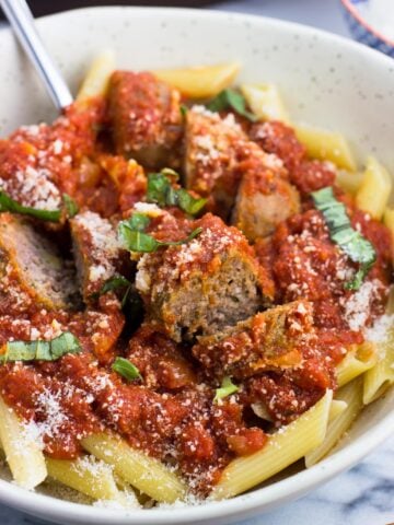 A bowl of Sunday sauce with meatballs and sausage served over penne pasta and garnished with Parmesan