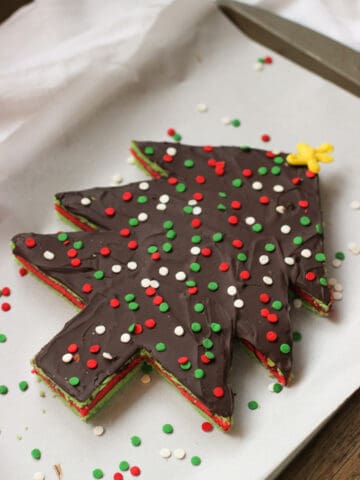 A Christmas tree-shaped and decorated slab of rainbow cookies on a parchment-lined baking sheet.