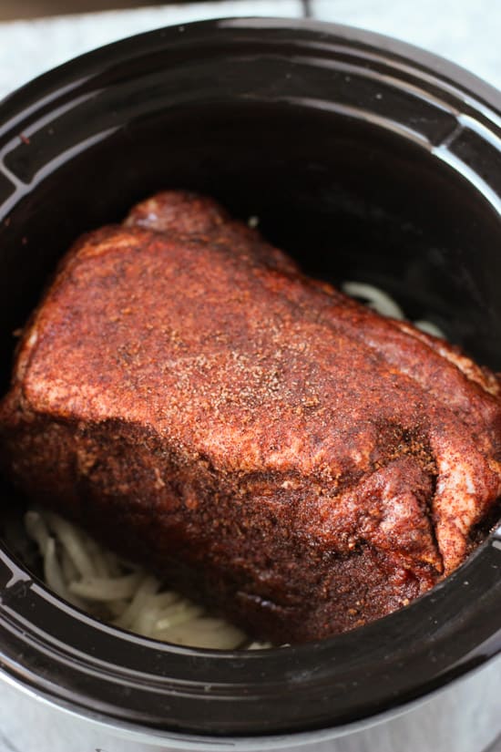 A spice-rubbed Boston butt pork roast in a round slow cooker set over sliced onion before cooking.