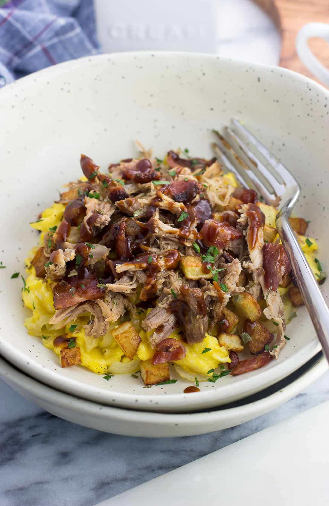 A pile of scrambled eggs, bacon, potatoes, pulled pork, and BBQ sauce in a bowl