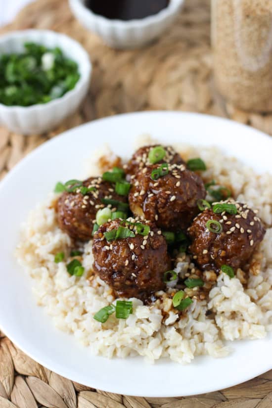 A plate of brown rice topped with hoisin meatballs covered in glaze, sesame seeds, and green onion