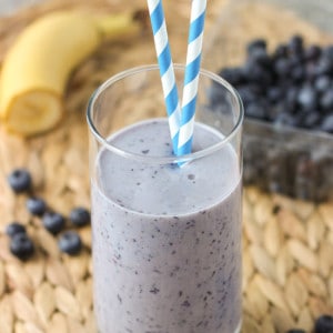 A smoothie in a tall glass with two paper straws surrounded by ingredients.