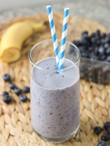 A smoothie in a tall glass with two paper straws surrounded by ingredients.