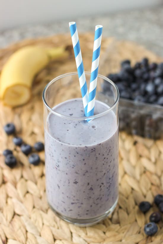 A smoothie in a tall glass with two paper straws surrounded by ingredients.