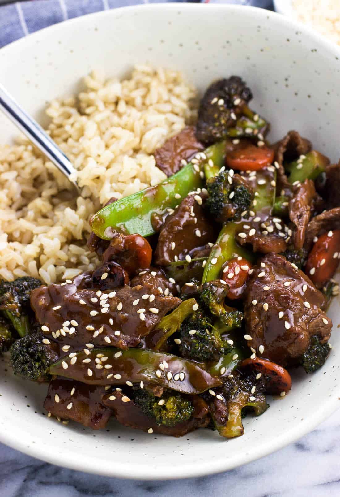 Beef and mixed vegetable stir fry in a bowl served beside brown rice.