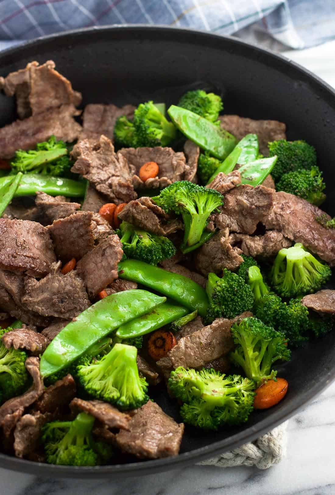 Cooked flank steak strips, broccoli florets, snow peas, and carrots in a skillet.