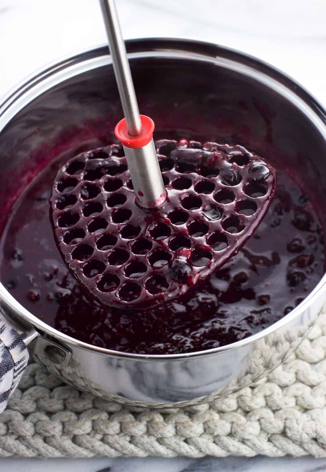 A potato masher being lifted out of a saucepan of jam after mashing the blueberries