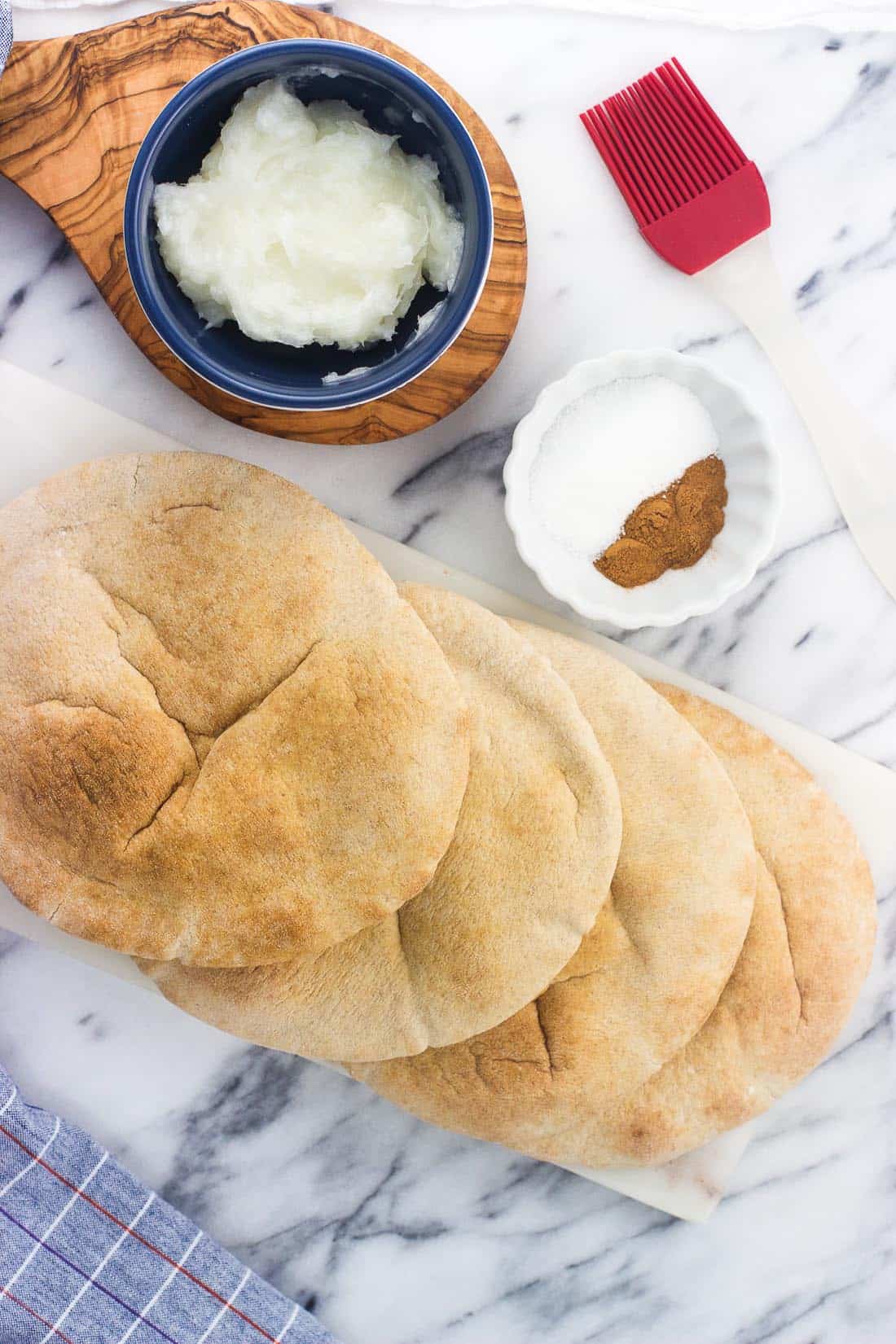 An overhead shot of whole wheat pitas, cinnamon, sugar, and coconut oil on a marble board