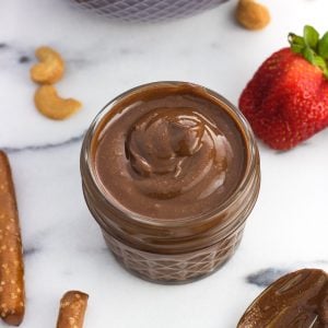 A small glass jar filled with creamy cashew butter next to a bowl of strawberries and pretzel rods