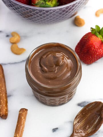 A small glass jar filled with creamy cashew butter next to a bowl of strawberries and pretzel rods