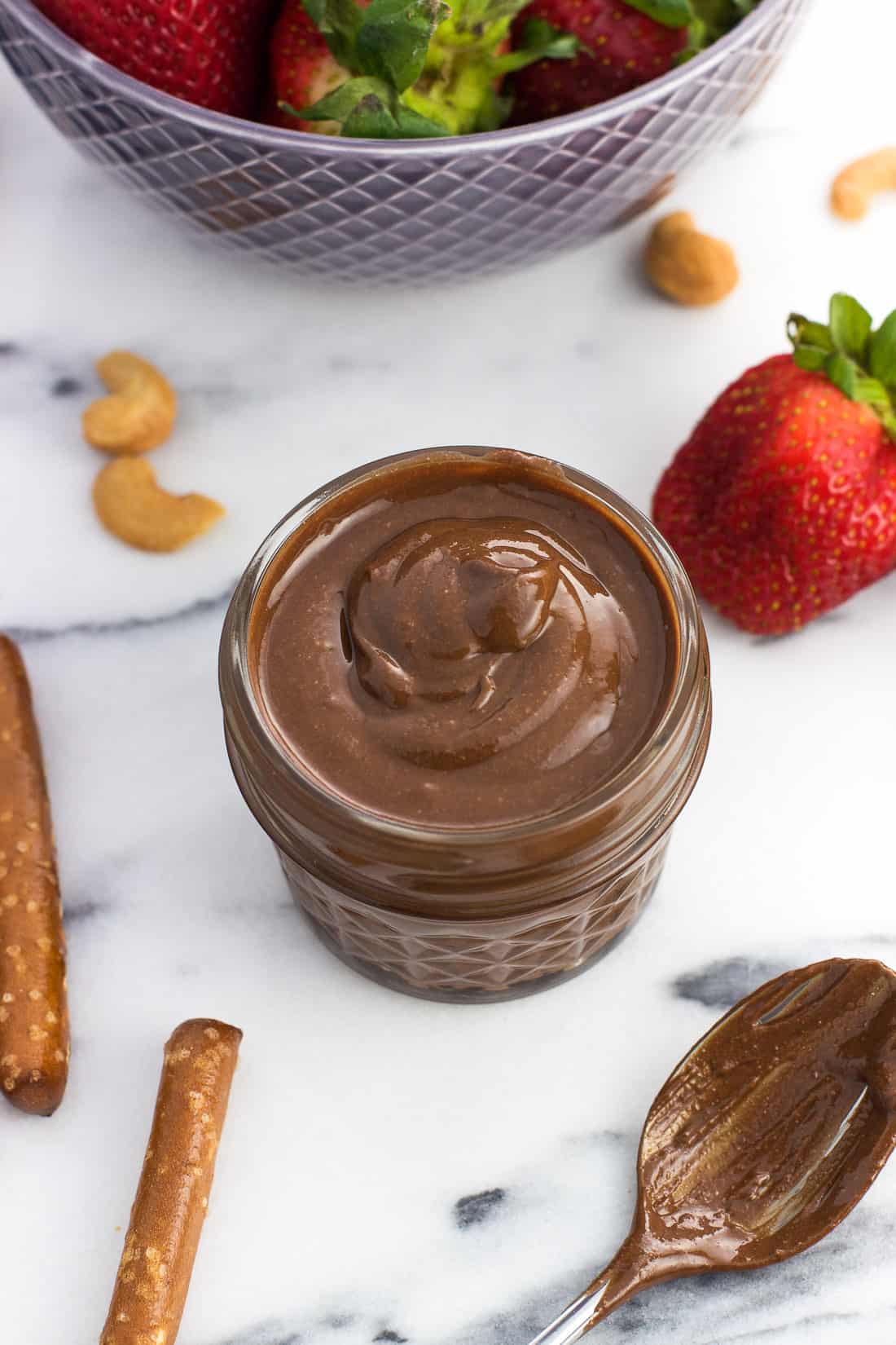 A small glass jar filled with cashew butter next to a bowl of strawberries and pretzel sticks