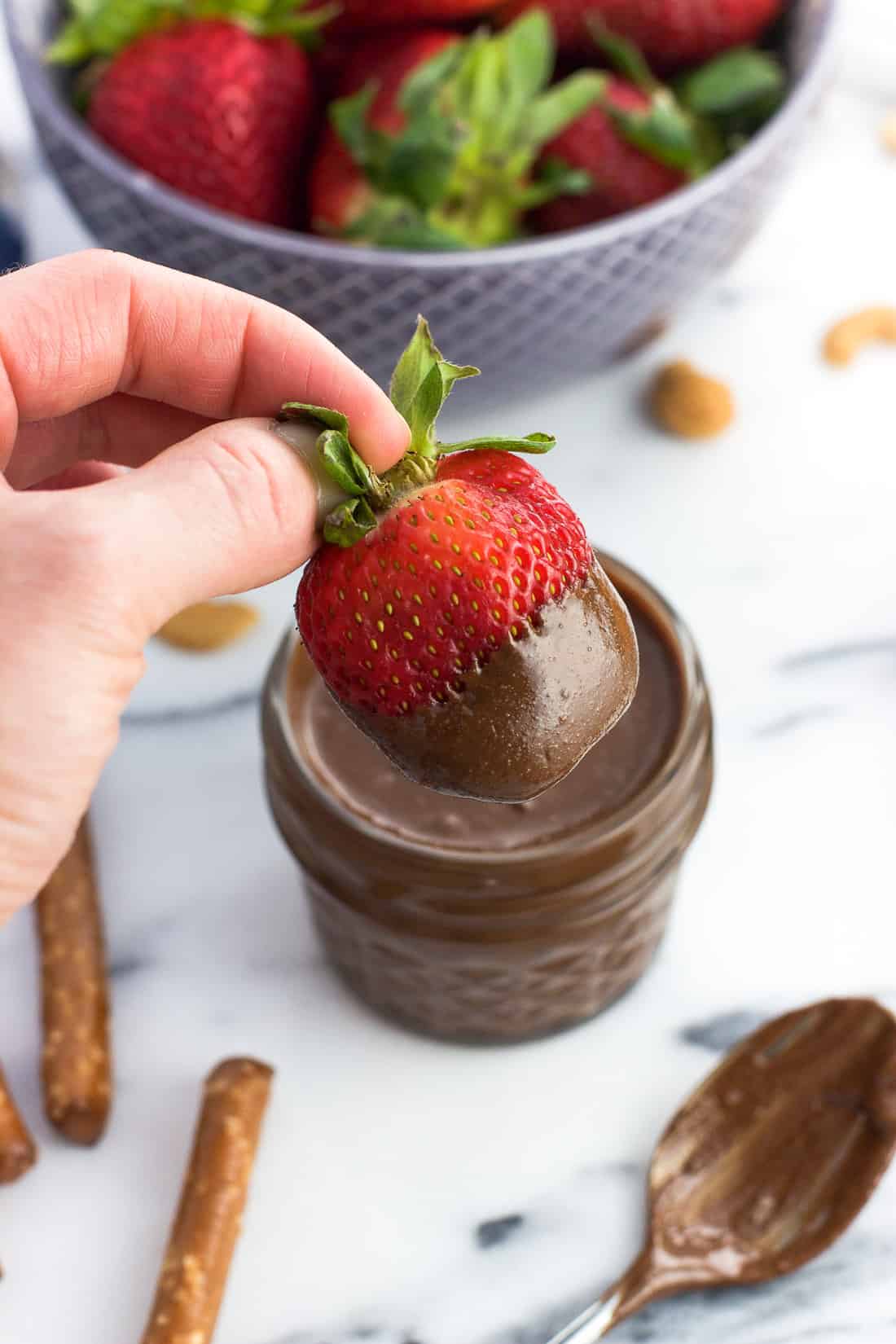 A hand holding up a fresh strawberry half dipped in the cashew butter