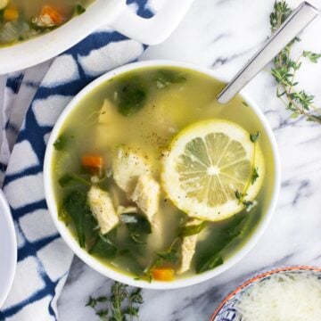 An overhead shot of a bowl of lemon chicken quinoa soup garnished with a lemon round next to a small bowl of grated Parmesan and a pepper mill