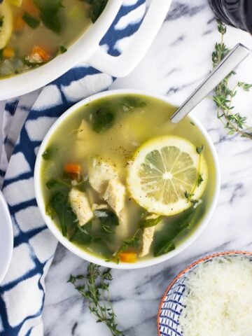 An overhead shot of a bowl of lemon chicken quinoa soup garnished with a lemon round next to a small bowl of grated Parmesan and a pepper mill