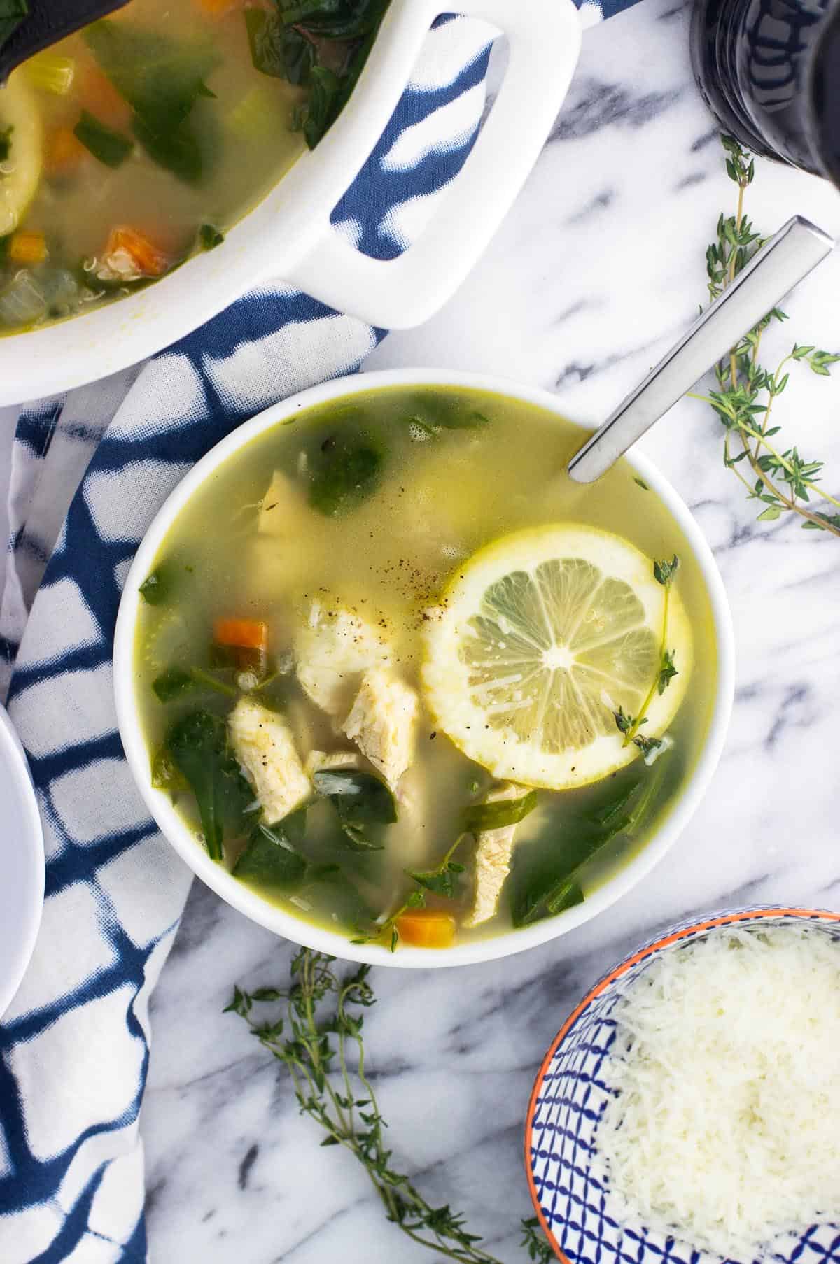 An overhead shot of a bowl of lemon chicken quinoa soup garnished with a lemon round next to a small bowl of grated Parmesan and a pepper mill.