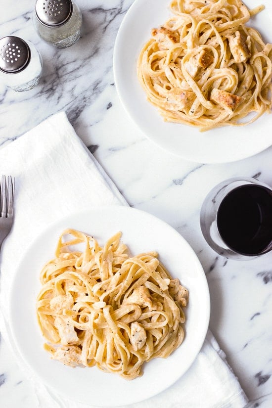Two plates of chicken and pasta on a table with a glass of wine and utensils.