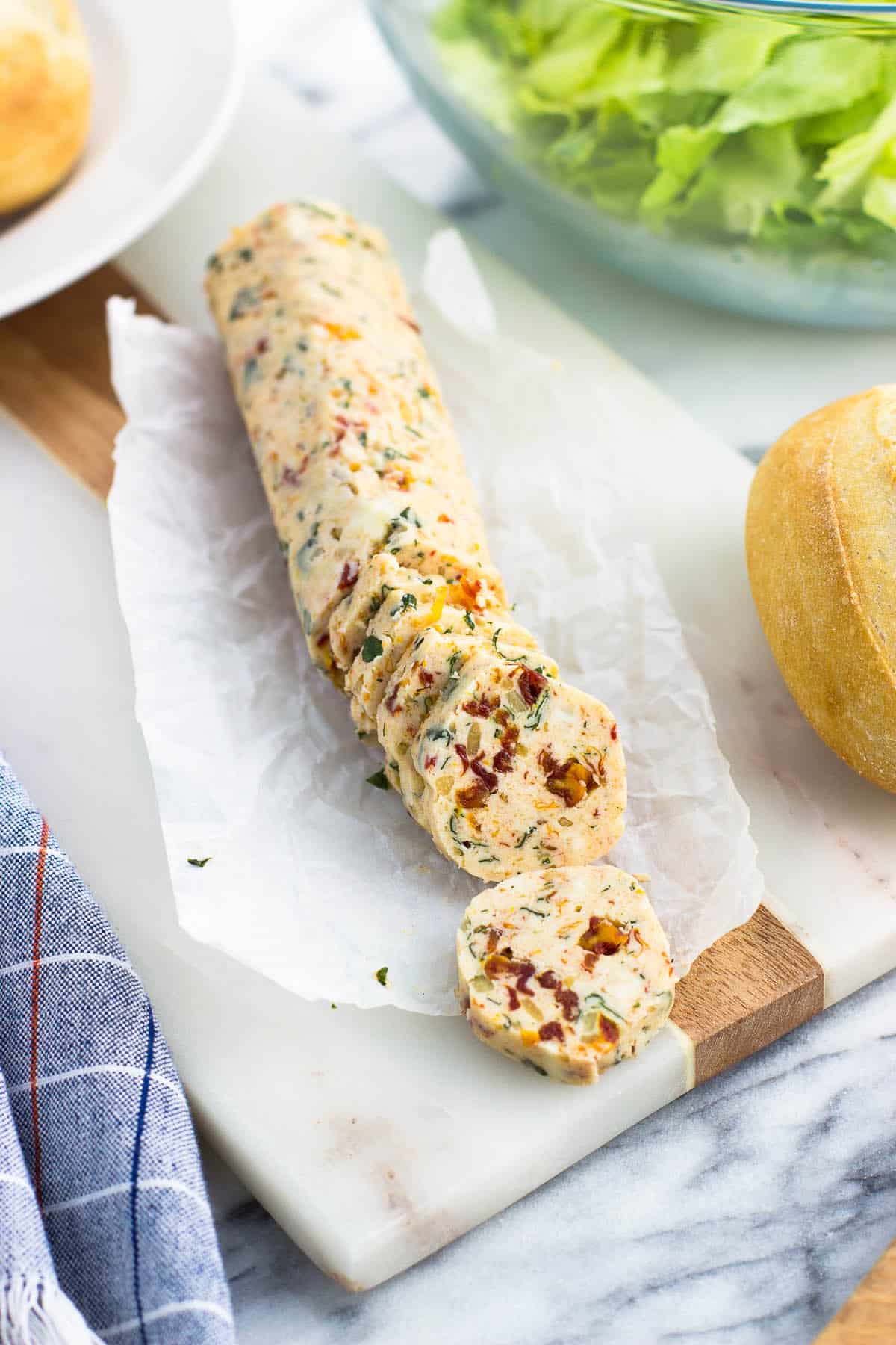 A chilled log of compound butter on a sheet of parchment paper next to a dinner roll and a bowl of salad